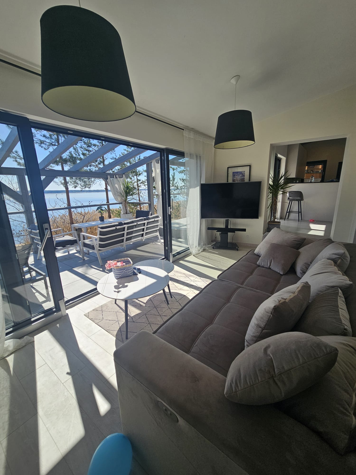 The image depicts a modern living room with a large window overlooking a patio and outdoor furniture, featuring a gray cou...