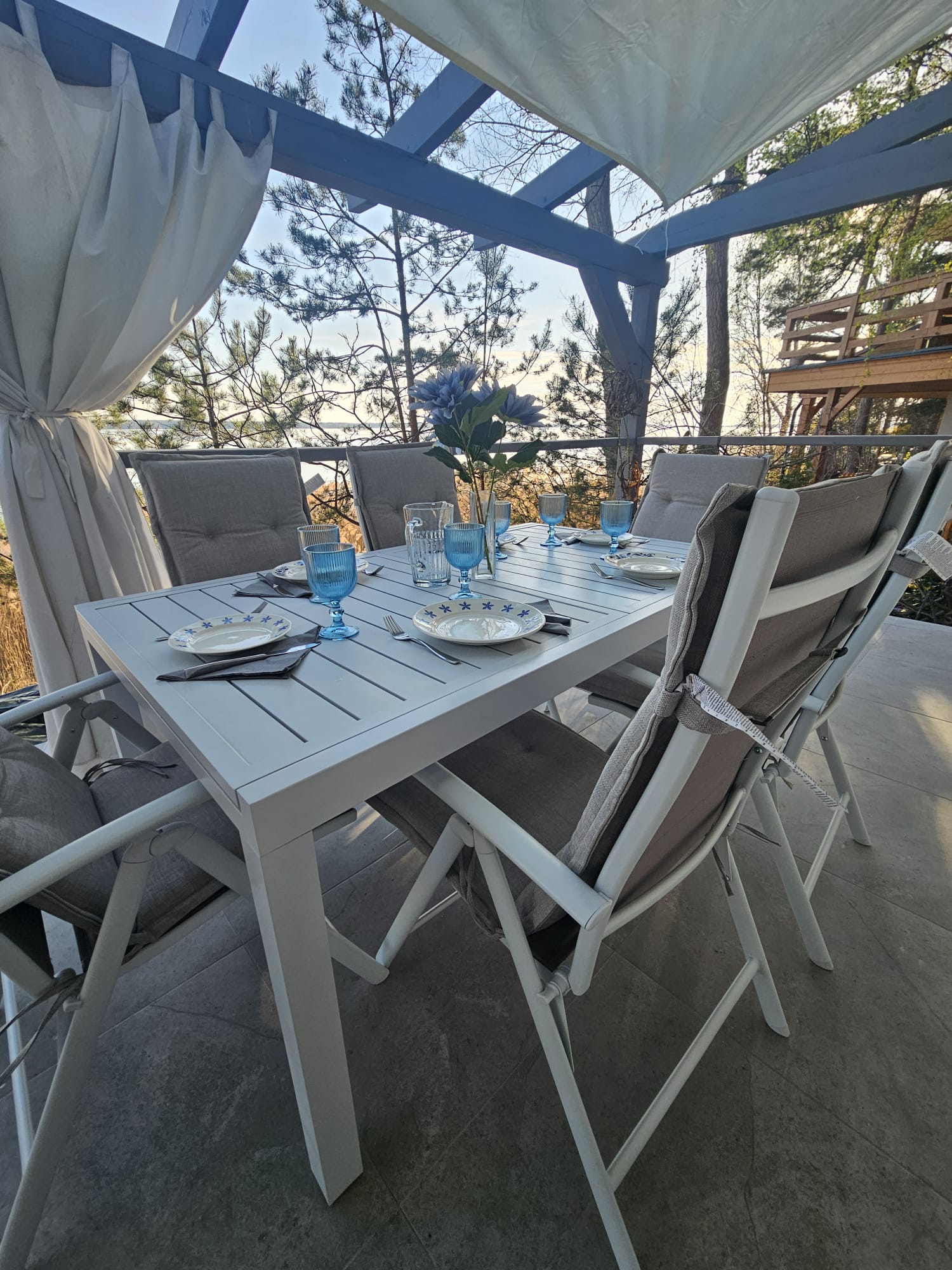 A dining table with six chairs set for a meal on a patio with trees and a body of water in the background.
