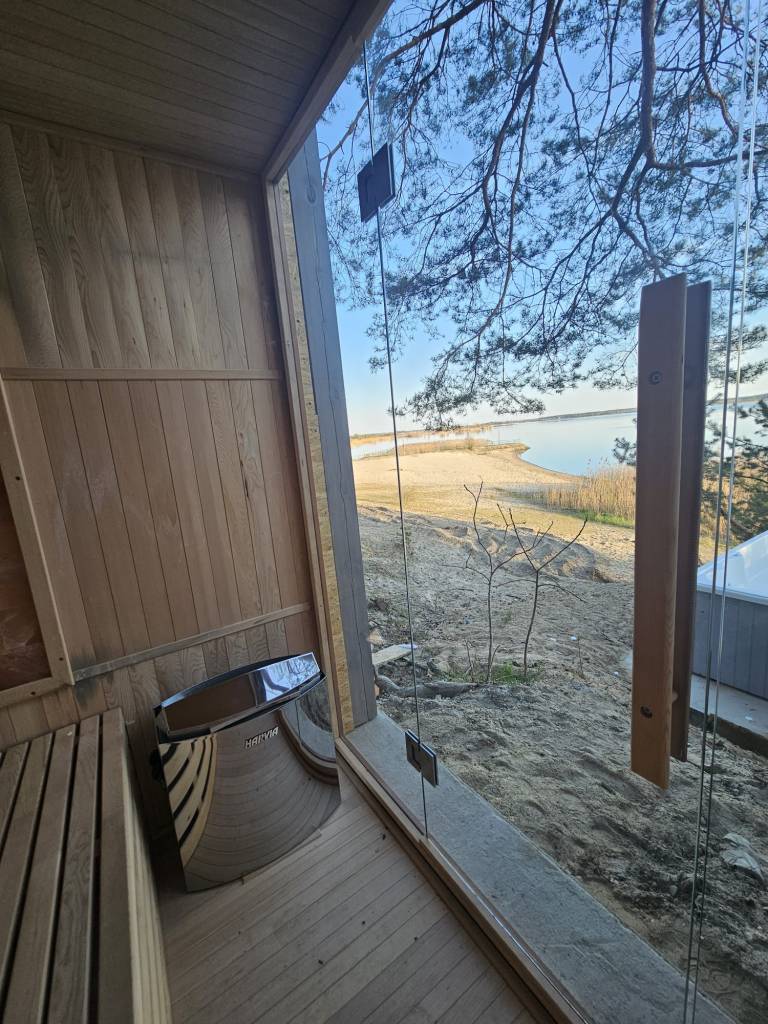 The image depicts a serene outdoor sauna situated on the edge of a lake, with a glass door and wooden walls that blend har...